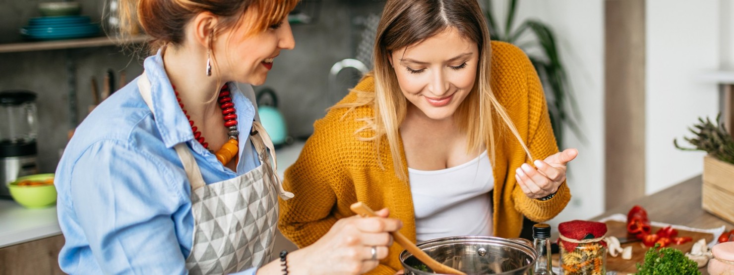 Eine circa 25-jährige, schwangere Frau kocht gemeinsam mit ihrer Freundin. Sie beugt sich über den Topf und genießt den Duft. Ihre Freundin rührt das Essen. Auf der Kücheninsel sind frisches Gemüse und Kräuter sowie ein Glas mit bunten Nudeln zu sehen.