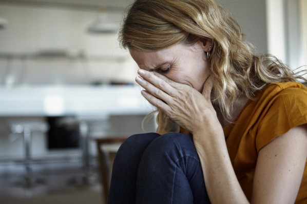 Ein Frau Mitte 40 schlägt traurig ihre Hände vor ihr Gesicht. Im Hintergrund erkennt man die Küche.