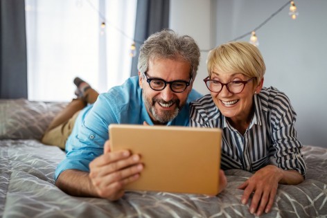 Ein etwa 55 jähriges Pärchen liegt auf einem Bett. Sie schauen gemeinsam auf ein Tablet und lachen.