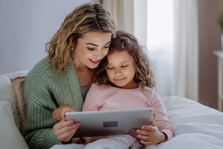 A happy mother with her little daughter lying on bed and using tablet at home.