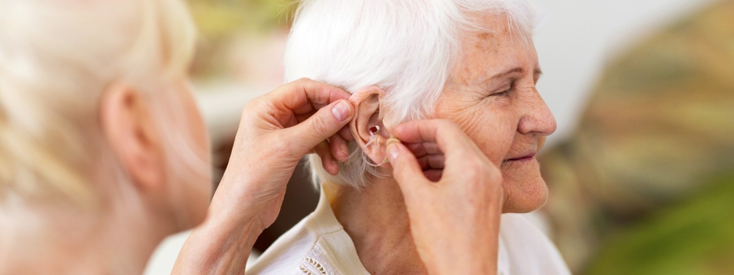 Einer Seniorin wird durch eine weibliche Person eine Hörhilfe in das Ohr eingesetzt.
