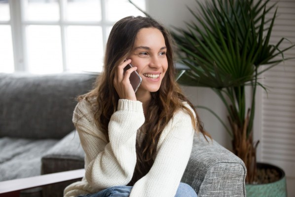 Eine etwa 25 jährige Frau sitzt auf einem Sofa und telefoniert. Dabei lächelt sie.