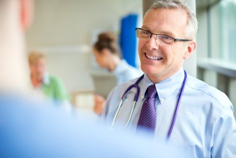 Ein Arzt um die 50 mit Brille steht im Krankenhaus auf dem Flur und führt ein Gespräch mit einem Patienten.