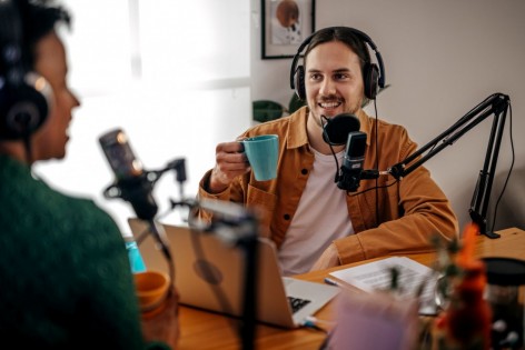 Zwei Männer sitzen sich an einem Tisch gegenüber und nehmen über Mikrofone eine Podcastfolge auf.
