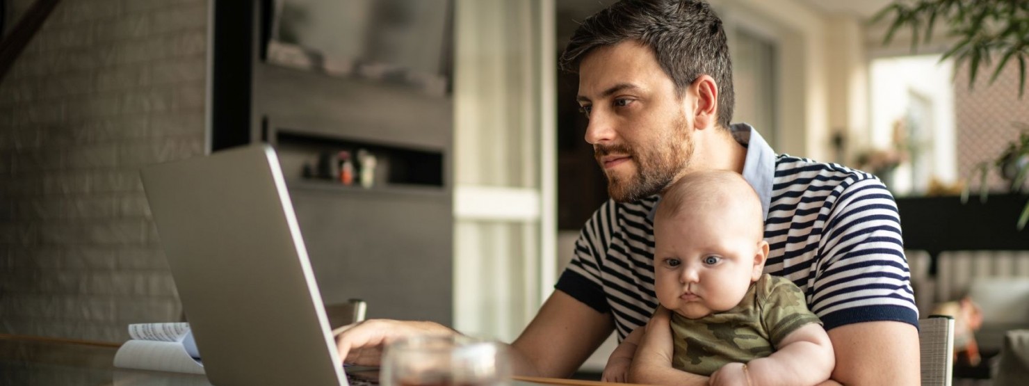 Ein etwa 35 jähriger Vater hat sein Baby auf dem Schoß und sitzt am Tisch- Er schaut interessiert auf seinen Laptop.