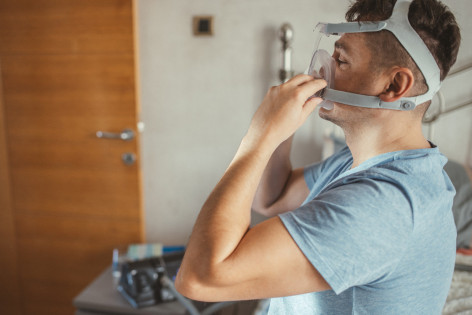 Ein Mann sitzt auf der Bettkante und trägt ein Beatmungsgerät.