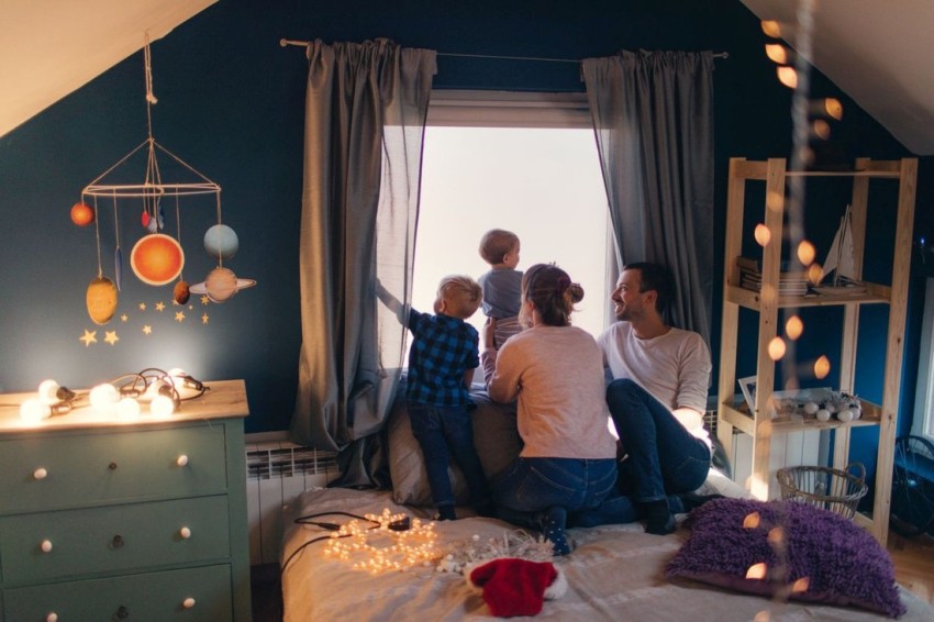 Junge Eltern sitzen mit ihren zwei Kindern am Fenster. Sie haben das Kinderzimmer mit Licherketten gemütlich geschmückt. 
