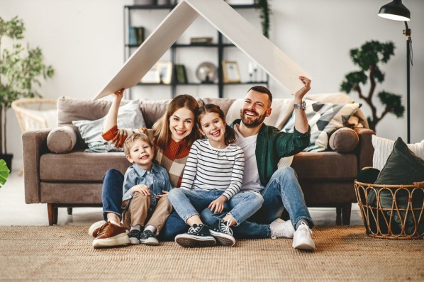 eine vierköpfige Familie sitzt in ihrem Wohnzimmer und hält ein Dach aus Pappe über ihre Köpfe