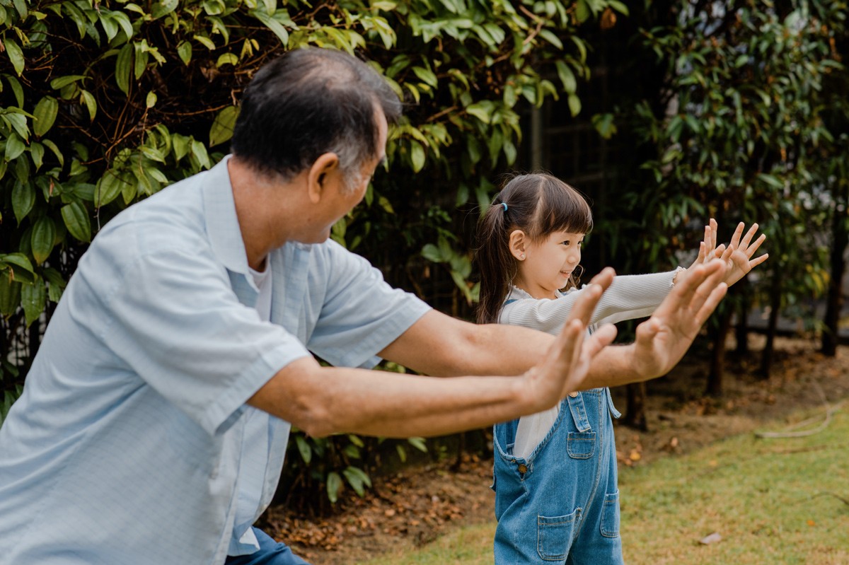 Ein etwa 70-jähriger Mann bringt seiner etwa 5-jährigen Enkelin Tai-Chi bei. Sie sind im Garten.