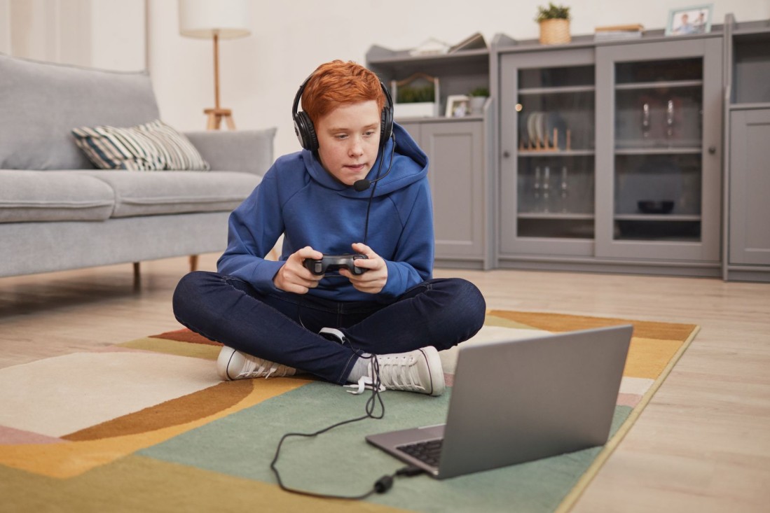Ein etwa 12-jähriger Junge sitzt auf dem Boden vor seinem Laptop und spielt mit Kopfhörern und dem Controller.