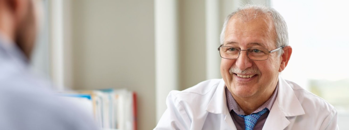Ein etwa 60 jährige Arzt sitzt an seinem Beratungstisch und berät einen Patienten. Dabei lächelt er.