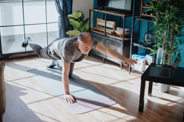 Ein etwa 65 jähriger Mann mach eine sportliche Übung in seinem Wohnzimmer auf einer Matte. Dabei streckt er einen Arm und ein Bein aus. Er schaut parallel auf einen Laptop.