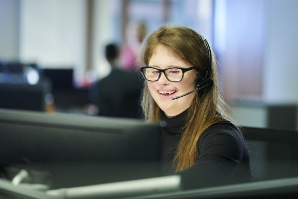 Eine etwa 25 jährige Frau mit Down-Syndrom sitzt auf der Arbeit an einem Computer und trägt ein Headset.