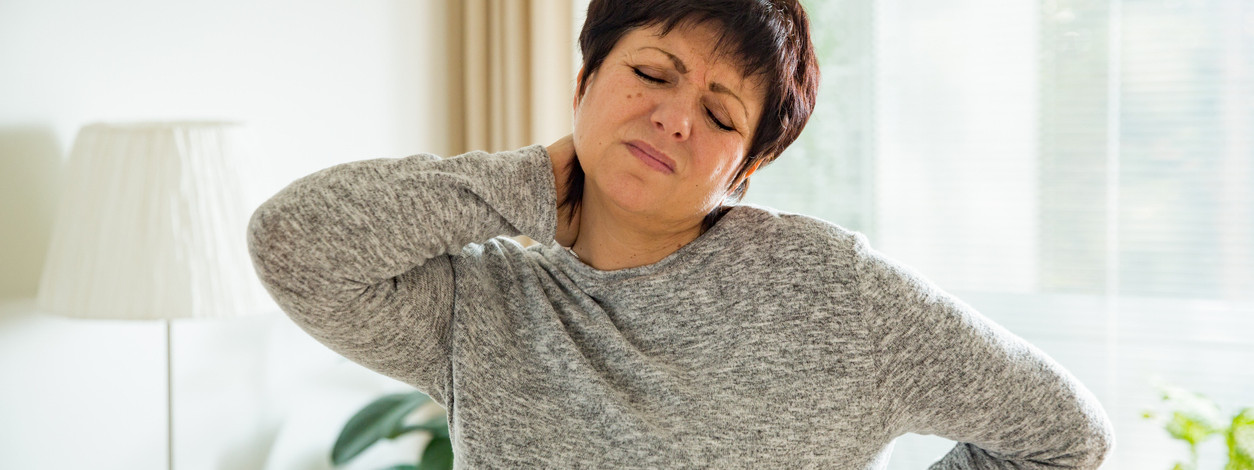 Mature woman suffering from backache at home. Massaging neck with hand, feeling exhausted, standing in living room.