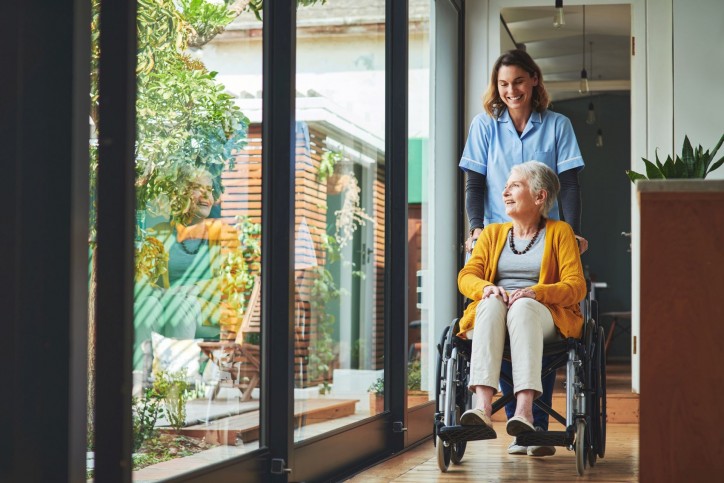 Eine Seniorin sitzt in einem Rollstuhl und wird von einer Pflegerin geschoben.