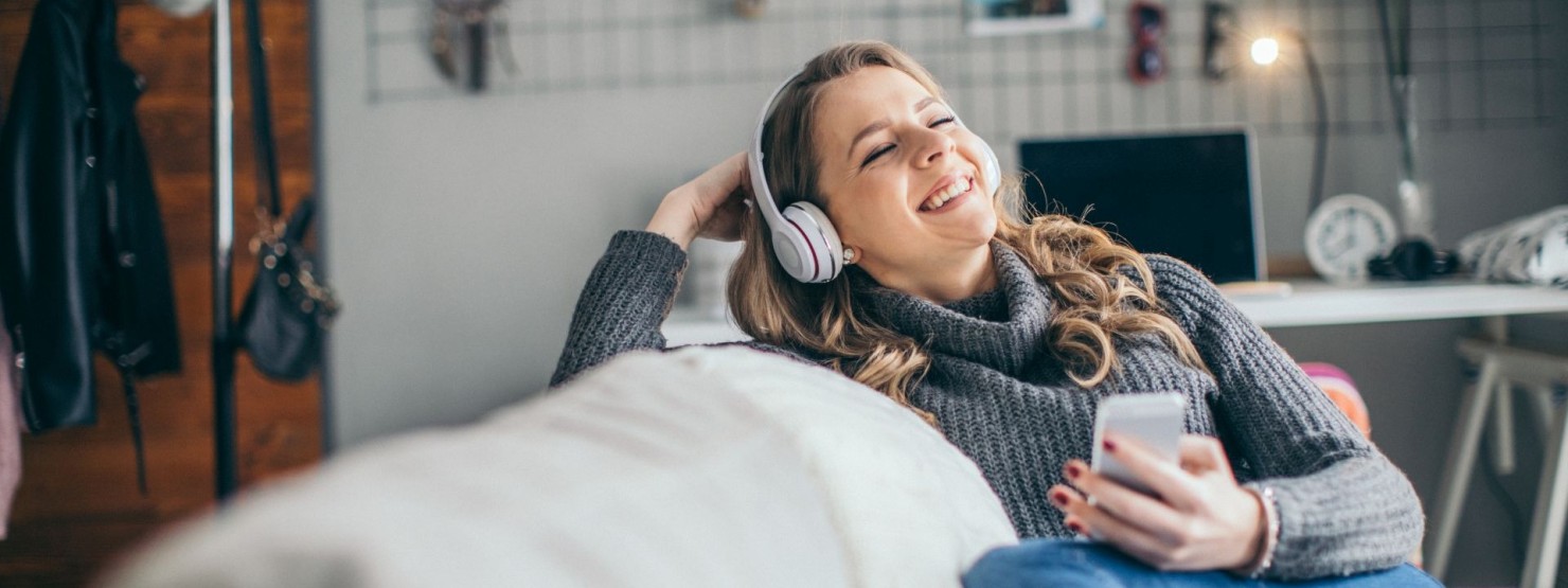 Eine etwa 30-jährige Frau sitzt auf dem Sofa und hört über Kopfhörer einen Podcast an.