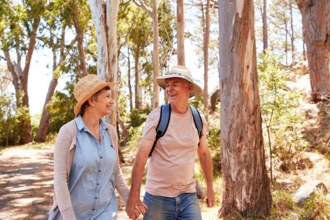 Ein etwa 55 jähriges Paar läuft hand ind Hand durch einen Wald. Beide lächeln sich an.