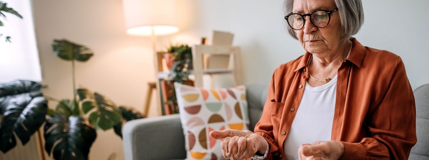 Einer Seniorin sitzt auf einem Sofa und ballt ihre Hände. Dabei schaut sie besorgt.