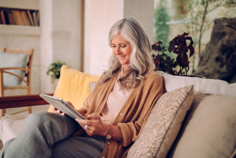 Eine Seniorin sitzt auf ihrem Sofa und schaut glücklich auf ihre Tablet.
