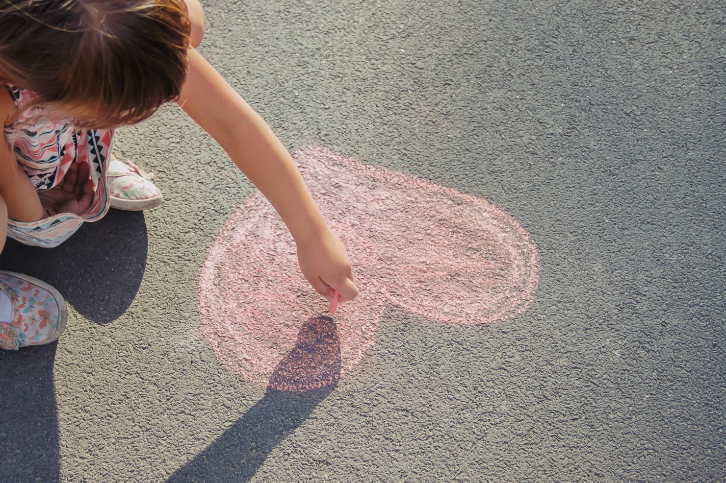 Ein kleines Mädchen, circa 7 Jahre alt, malt mit rosa Kreide ein Herz auf den Asphalt.