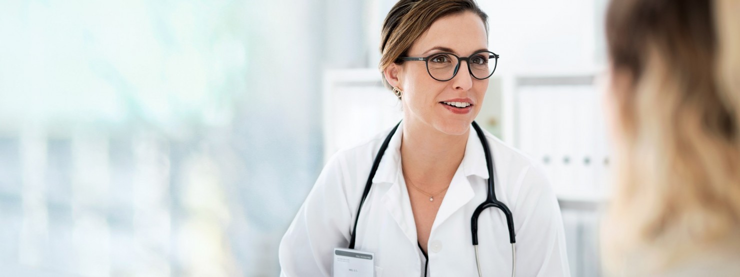 Eine Ärztin sitzt an einem Tisch und berät einen Patienten. Auf dem Tisch stehen ein Laptop und eine Packung Tabletten.