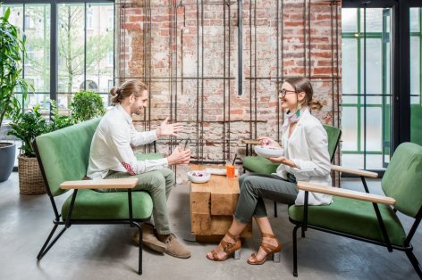 Zwei etwa 25 jährige Personen sitzen jeweils auf zwei Sofas in einem Café. Beide unterhalten sich und essen etwas aus einer Schale.