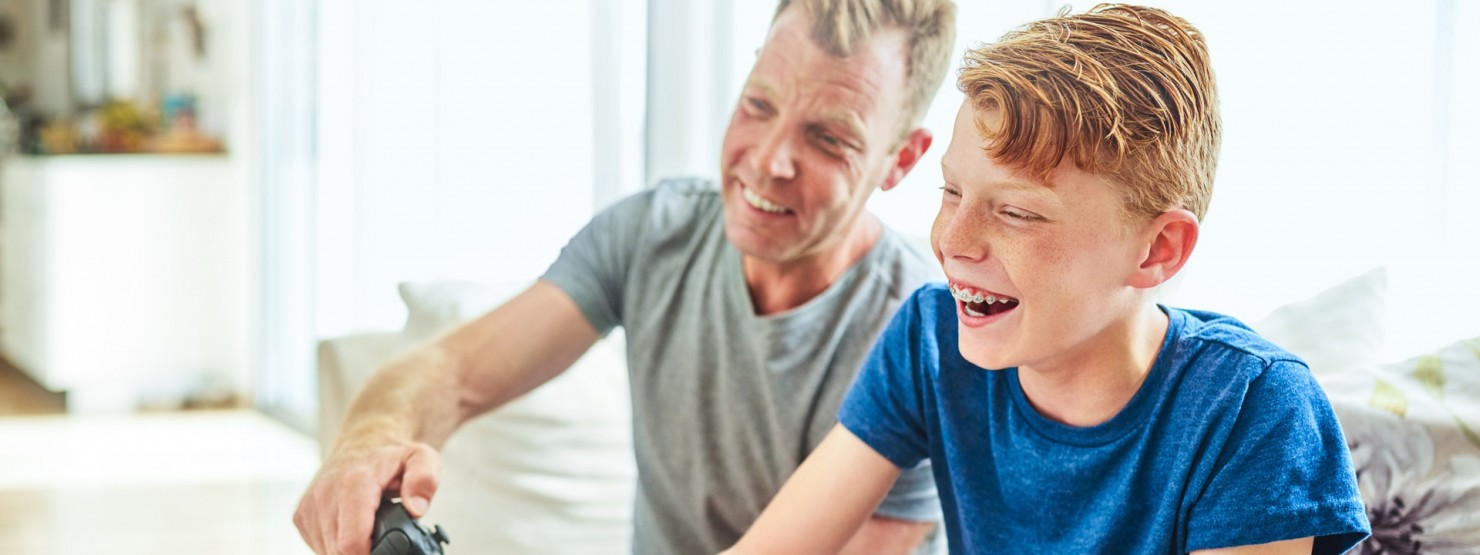 Ein Junge mit blauen Tshirt und Zahnspange sitzt mit einem erwachsenen Mann auf dem Sofa und spielt auf der Spielkonsole. 