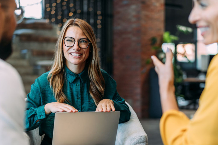 Eine Beschäftigte spricht fröhlich mit ihren Kollegen. Dabei hat sie ihren Laptop auf dem Schoß.