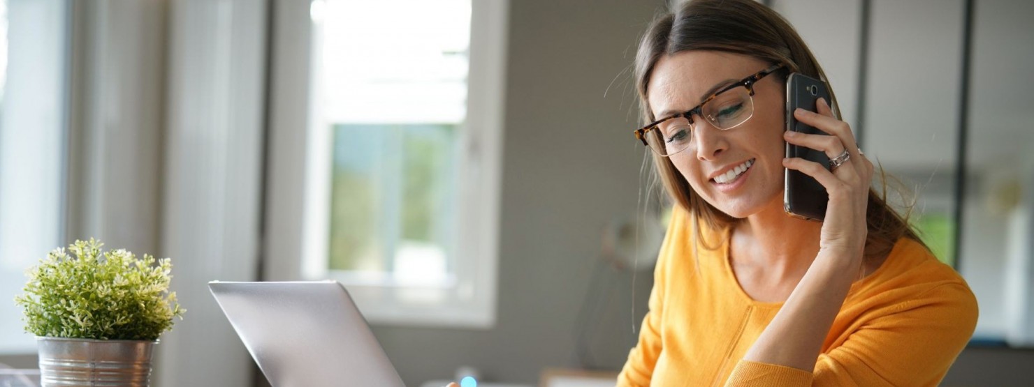 Eine etwa 40 jährige Frau sitzt an einem Schreibtisch. Sie arbeitet an einen Laptop und telefoniert parallel. Dabei lächelt sie.