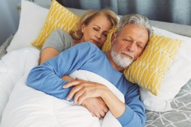 Ein Paar, Ende 50 liegt schlafend im Bett. Die Frau liegt hinter dem Mann und umfasst seinen Körper.