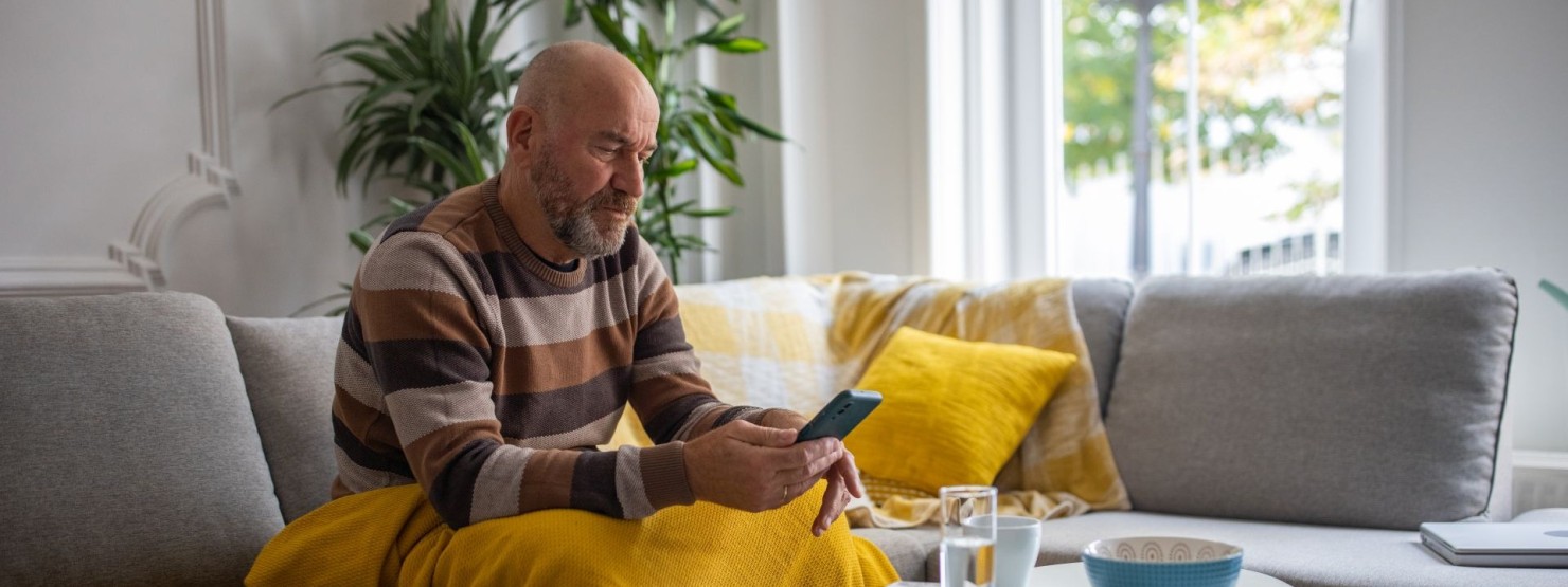 Ein etwa 55-jähriger Mann sitzt auf dem Sofa und schaut auf sein Handy.