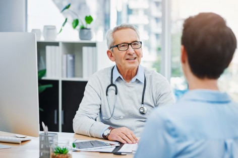 Ein etwa 60 jähriger Arzt sitzt an einem Schreibtisch und berät einen etwas 30 jährigen Patienten.