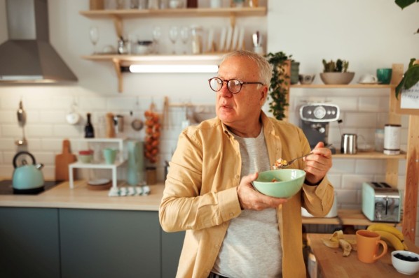 Ein etwa65-jähriger Mann isst stehend Müsli in der Küche.