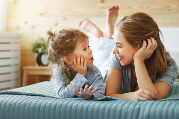 Eine Mutter liegt mit ihrer etwa 5 jährigen Tochter auf einem Bett. beide sprechen miteinander und lächeln.