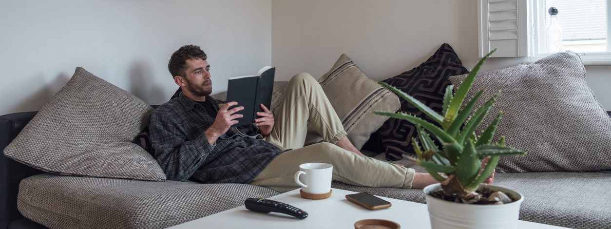A caucasian man wearing casual clothing, sitting on a sofa in his apartment and reading a book.