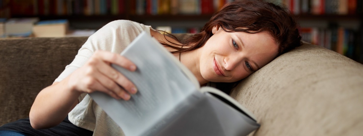 Eine etwa 30-jährige Frau liest mit lächelndem Gesicht ein Buch auf der Couch. Ihr Kopf liegt entspannt auf der Rückenlehne.