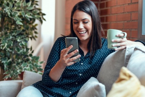Eine Frau sitzt auf einem Sofa, hält eine Tasse in linken und in der rechten Hand ein Handy.