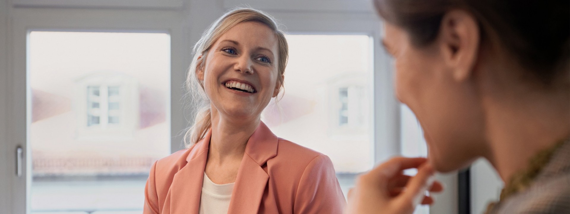 Eine etwa 30 Jährige Frau mit langen blonden Haaren und einem lachsfarbenem Jacket lächelt eine zweite unscharfe Person an. Diese steht die im Vordergrund rechts. Sie befinden sich in einem Büro.