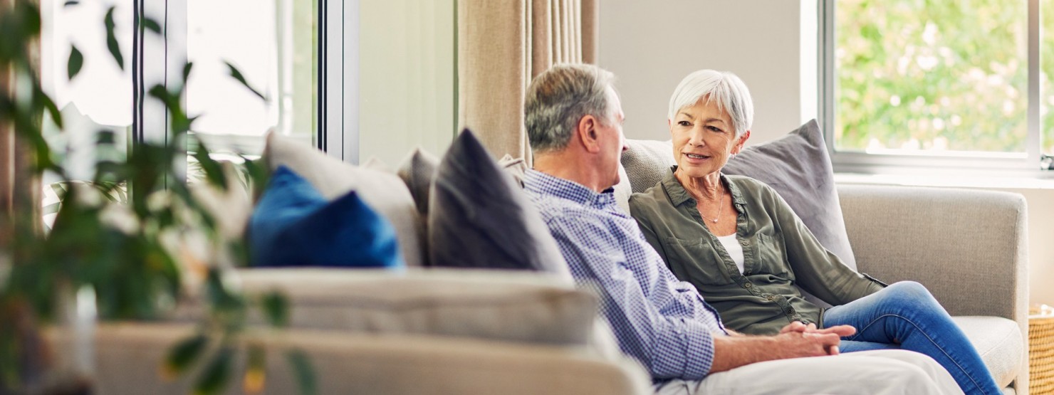 Eine Frau und ein Mann, beide etwa 60 Jahre alt, unterhalten sich auf einem Sofa.