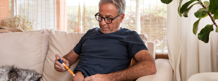 Ein etwa 55-jähriger Mann sitzt auf dem Sofa im Wohnzimmer und spritzt sich mit einem Fertigpen in die Bauchfalte.