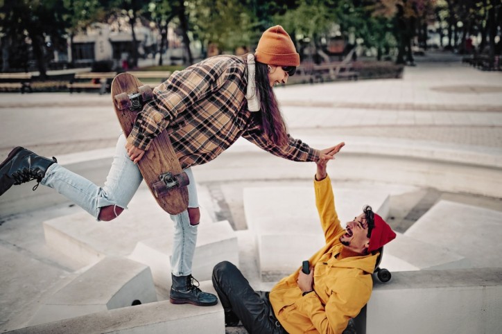 Eine junge Frau und ein junger Mann, beide Mitte 20, sind zusammen auf einem gepflasterten Platz. Er sitzt zwischen zwei Steinen einer dekorativen Steinformation angelehnt an ein Skateboard. Sie steht auf einem der Steine und hält ein Skateboard unterm Arm. Sie klatschen einander ausgelassen ab. Sie tragen herbstliche Kleidung und Mützen.