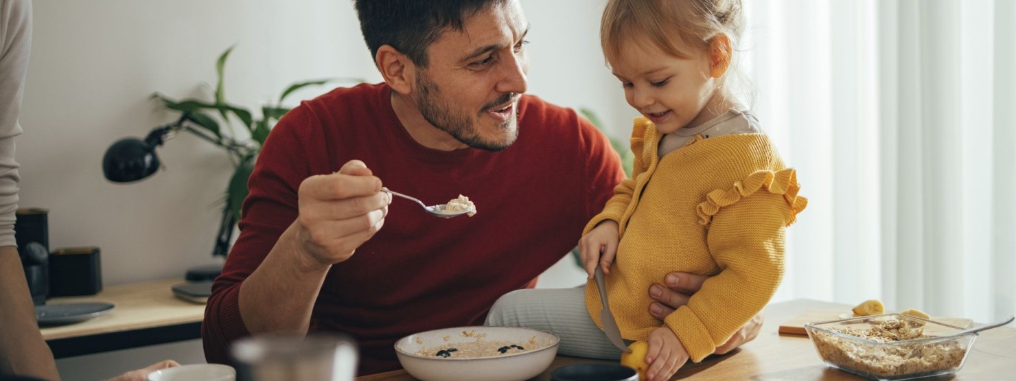 Ein Vater sitzt mit seiner kleinen Tochter am Esstisch. Er füttert das Mädchen mit Müsli.