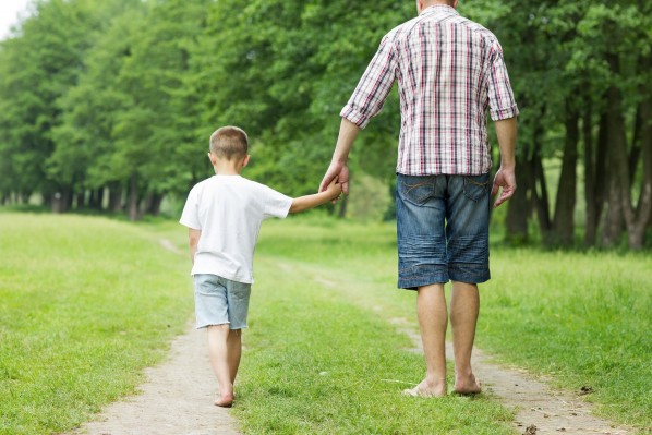 Ein Vater läuft mit seinem Sohn einen Feldweg an einer Wiese entlang. Beide fassen sich an den Händen. Sie laufen barfuß.