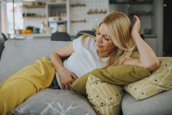 Eine etwa 25 jährige Frau liegt auf einem Sofa. Sie hält sich den Bauch und schaut schmerzverzehrt.