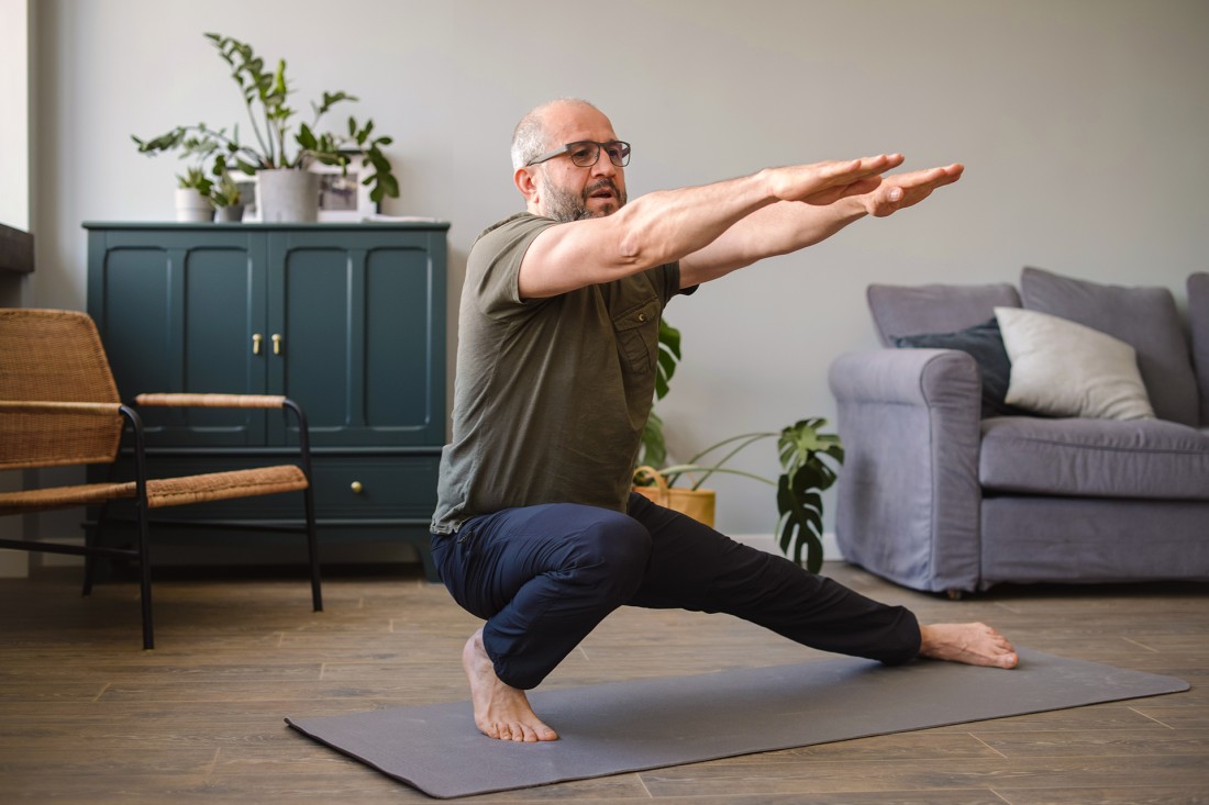 Ein etwa 50 Jahre alter Mann macht zu Hause im Wohnzimmer Yoga. Er Hockt auf einer Yogamatte und streckt die Arme aus.