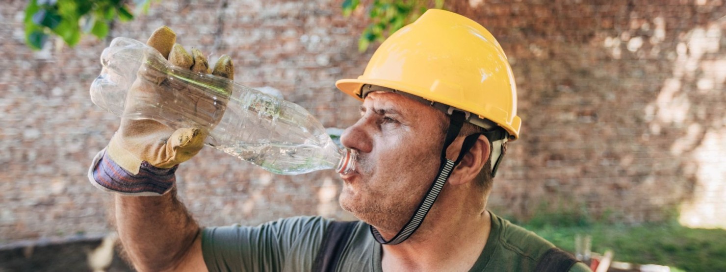 Ein Baumarbeiter schwitzt stark und trinkt dagegen Wasser aus einer Flasche.