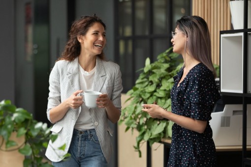 Zwei etwa 35 jährige Frauen stehe zusammen auf der Arbeit und unterhalten sich in ihrer Pause. Eine Frau hält eine Kaffeetasse in der Hand. Beide lächeln.