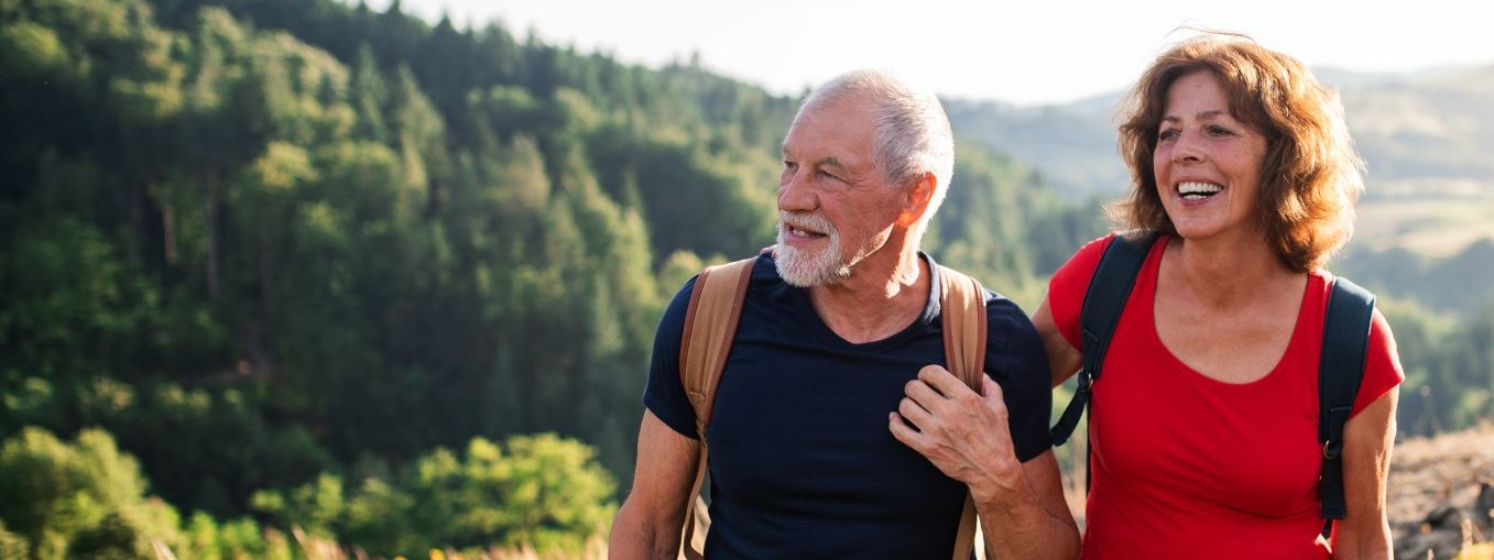 Ein etwa 60 jähriges Pärchen wandert durch hügelige Natur. Beide haben Rucksäcke auf, schauen in die gleiche Richtung und lächeln.