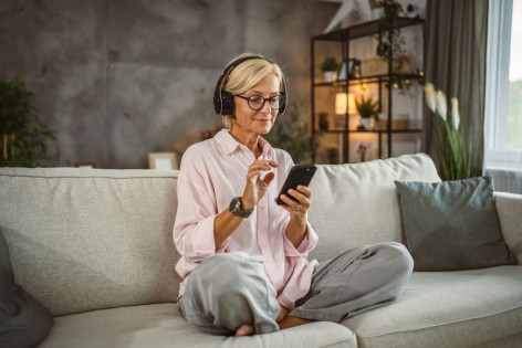 Eine Frau sitzt im Schneidersitz auf einer Coach und hört über Kopfhörer den AOK-Podcast zum Thema Pflege.