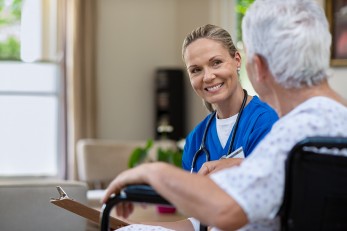 Eine blonde Frau mit blauem Kittel und Stethoskop um den Hals sitzt einer Seniorin gegenüber. Beide lächeln sich an. 